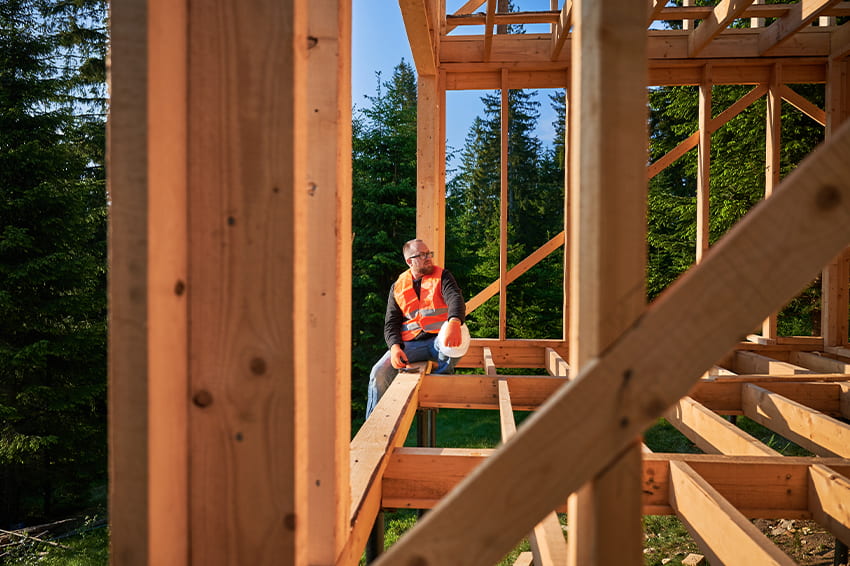 Cieśla w kasku na tle drewnianego szkieletu domu wykonanego z tarcicy iglastej w lesie.