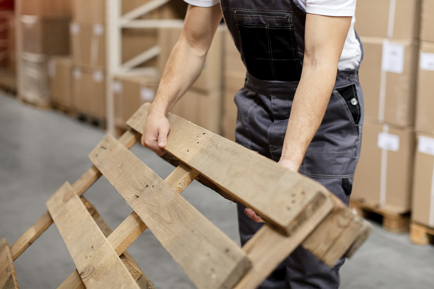 Pracownik magazynu w ubraniu roboczym trzymający surową deskę paletową na tle regałów z towarem.