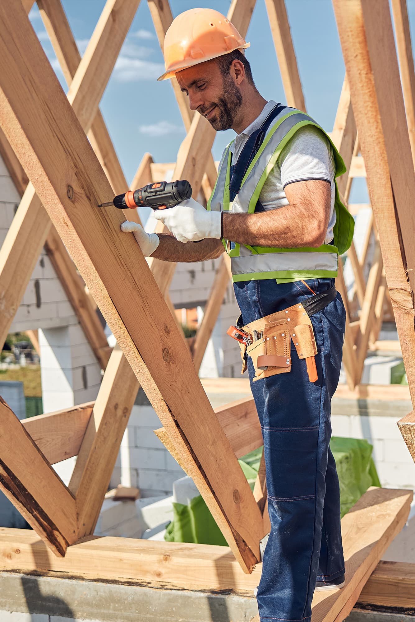 Cieśla w pomarańczowym kasku i kamizelce odblaskowej skręca wkrętarką elementy drewnianej więźby dachowej na budowie domu.