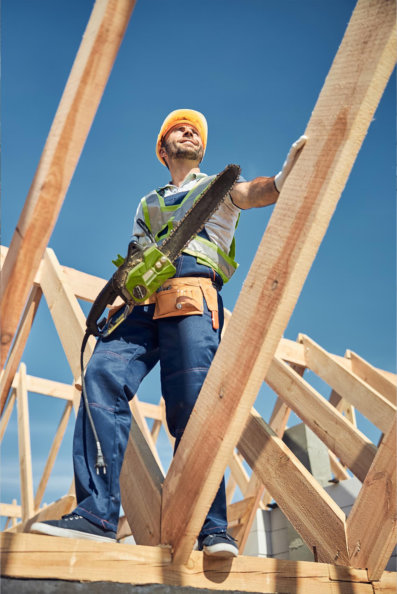 Cieśla w kasku ochronnym z piłą łańcuchową stojący na drewnianej konstrukcji więźby dachowej podczas prac montażowych.